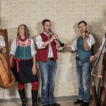 Fünf Personen der Musikgruppe Liadhaber stehen mit ihren Instrumenten nebeneinander in einer Reihe vor einer Wand in einem alten Gebäude