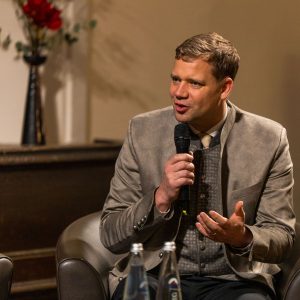 Mann im Trachtenanzug mit Mikrofon auf einem Sessel sitzend, sprechend. Erster Vorsitzender Dr. Olaf Heinrich