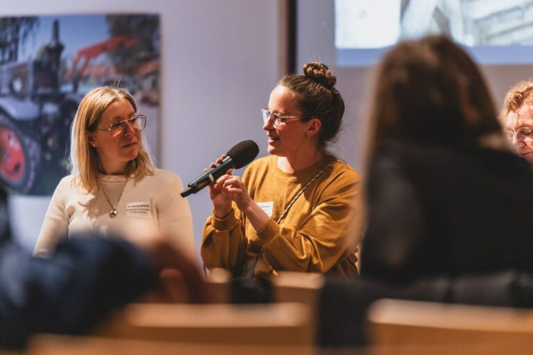 Zwei Frauen bei einem Gespräch in einer Tagungssituation, eine hält ein Mikrofon und spricht