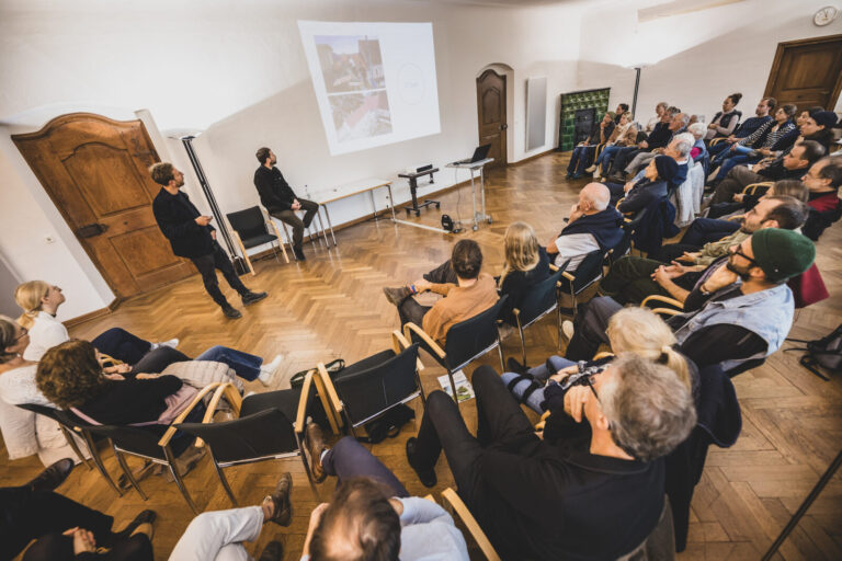 Eine große Gruppe von Menschen sitzt halbkreisförmig um zwei Frauen, die ein Präsentation halten. Die Menschen sitzen auf Stühlen in einem historischen Raum mit Holz-Parkett.
