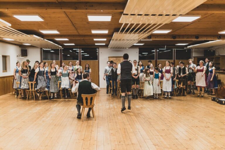 Eine große Gruppe junger Menschen in bayerischer Tracht schaut in Richtung Kamera und springt in die Höhe. Vor Ihnen stehen zwei Volksmusik-Referenten in Tracht, einer von Ihnen springt, der andere sitzt auf einem Stuhl mit einer Gitarre.