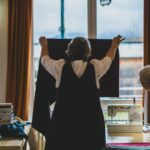 Zwei Frauen arbeiten an einem Kleidungsstück, die eine hält ein Textilstück vor ein Fenster, die andere schaut von der Seite darauf. Nähmaschinen sind auf tischen zu sehen.