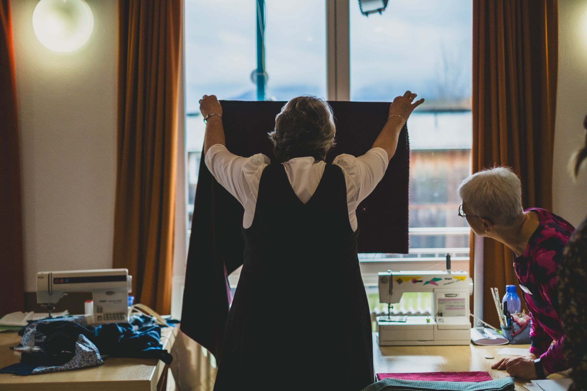 Zwei Frauen arbeiten an einem Kleidungsstück, die eine hält ein Textilstück vor ein Fenster, die andere schaut von der Seite darauf. Nähmaschinen sind auf tischen zu sehen.