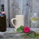 Flasche Bier, Steinkrug und Glas Wein vor einer Holzwand, im Bildvordergrund liegt noch eine Rose