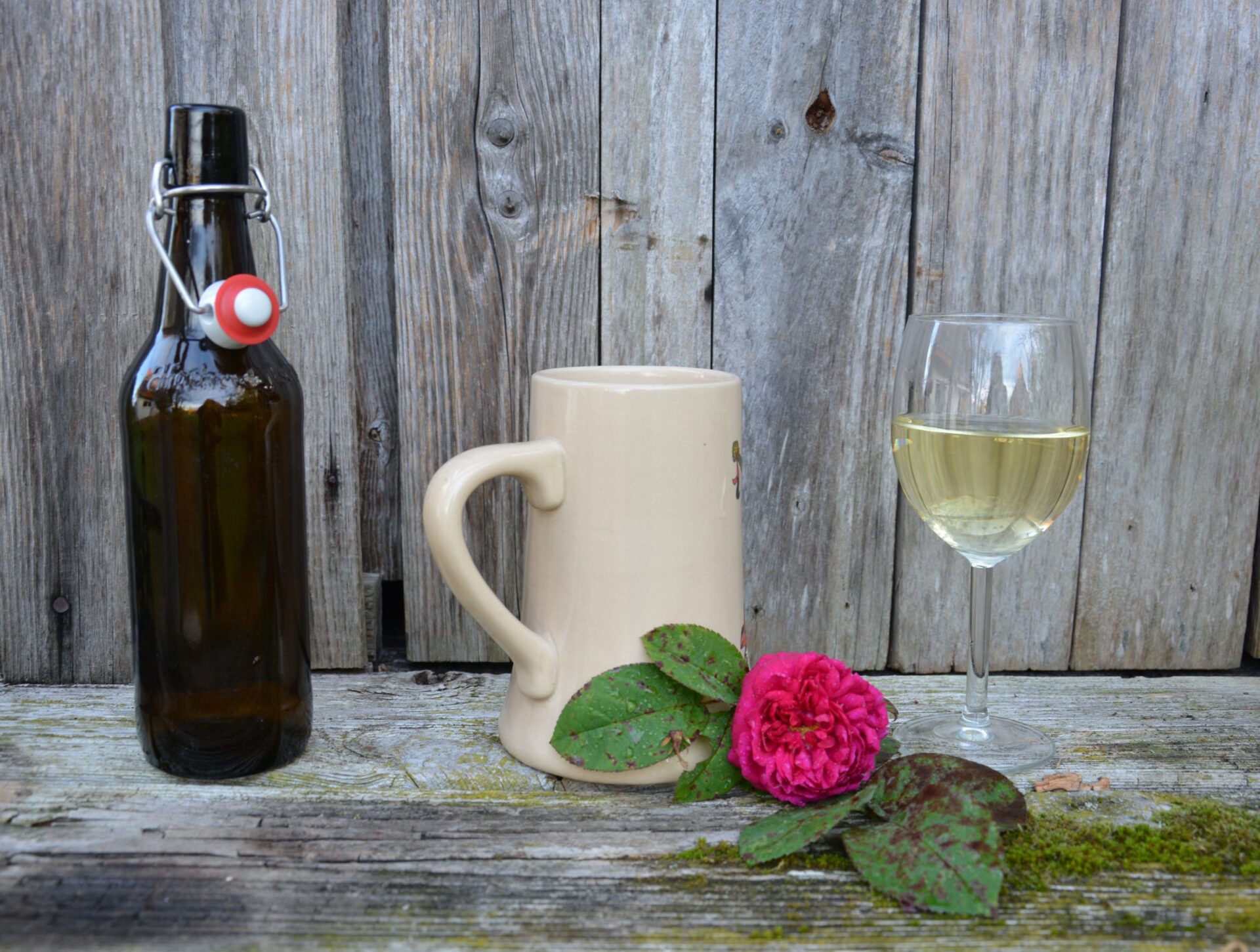 Flasche Bier, Steinkrug und Glas Wein vor einer Holzwand, im Bildvordergrund liegt noch eine Rose
