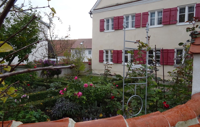 Ein altes Schulhaus mit roten Fensterläden, davor ein Garten mit blühenden Rosen