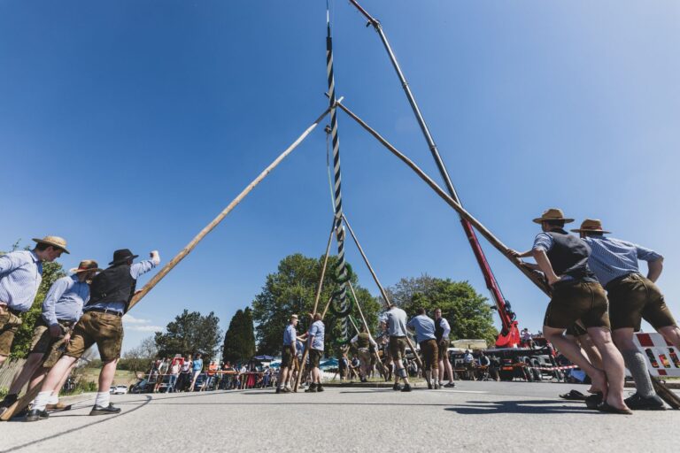 Männer in Tracht stellen auf einem Platz einen Weiß-Blau-gestreiften Maibaum mithilfe von langen Holzstöcken und einem Kran an. Der Himmel ist blau.