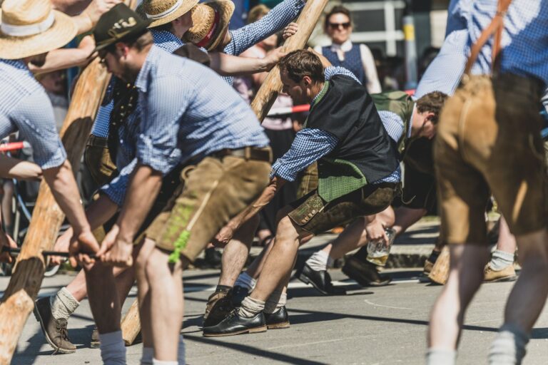 Männer in Tracht stellen auf einem Platz einen Weiß-Blau-gestreiften Maibaum auf.