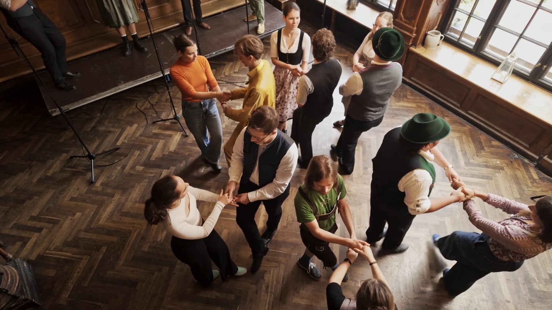 Menschen tanzen in Paaren Volkstänze auf einem Parkettboden, manche sind in Tracht. Die Perspektive ist von oben.